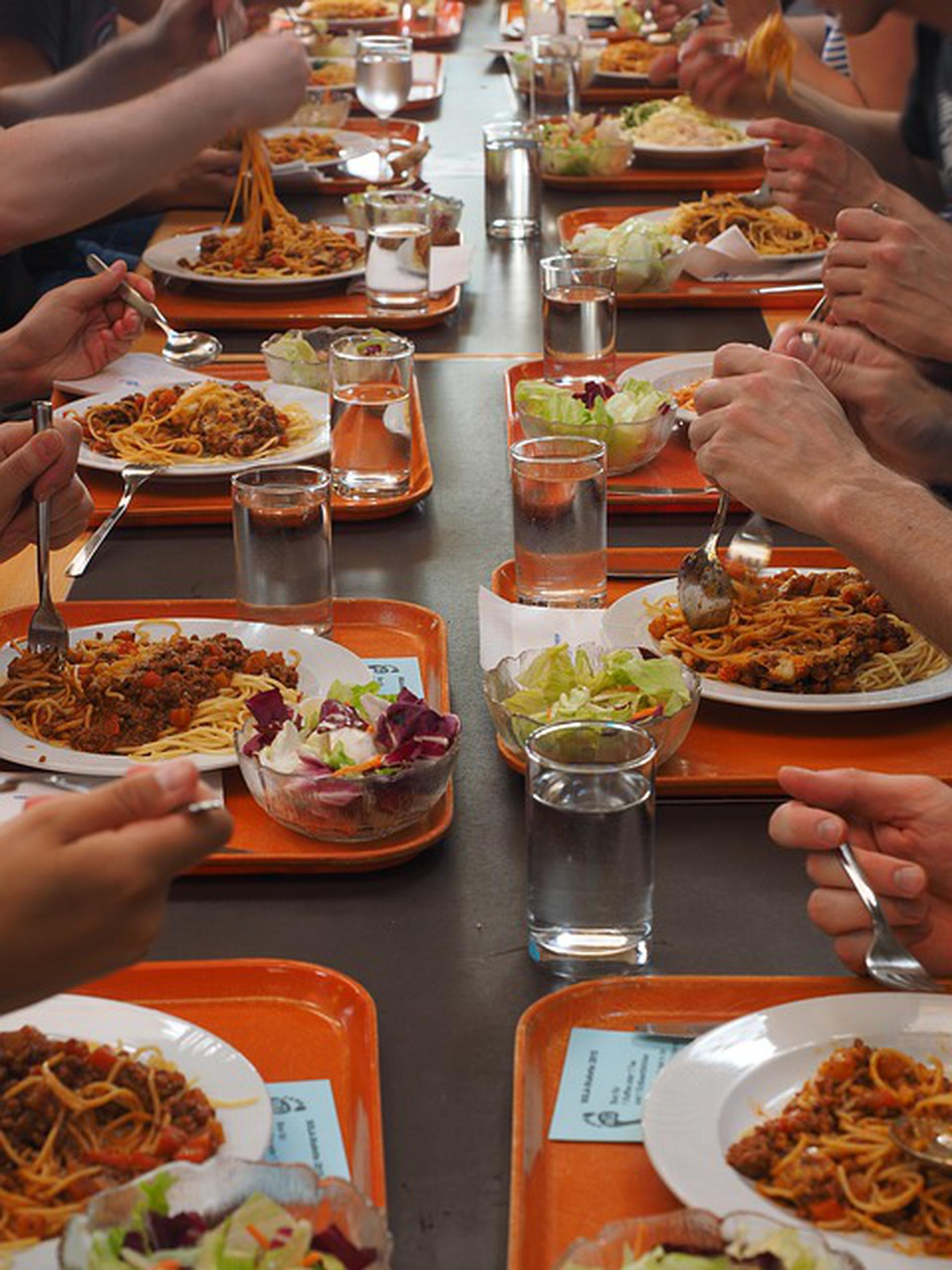 deux rangées d'enfants avec plateaux de repas sur tables longue mangent à la cantine